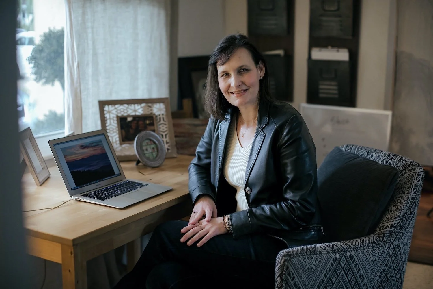A woman with dark hair, wearing a black leather jacket and a white top, smiling and sitting at a wooden desk with a laptop, framed photos, and decorative items in the background.