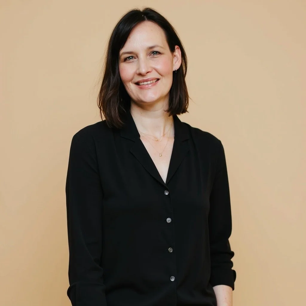 A woman with shoulder-length dark brown hair and light skin, smiling, wearing a black button-up shirt, standing against a beige background.