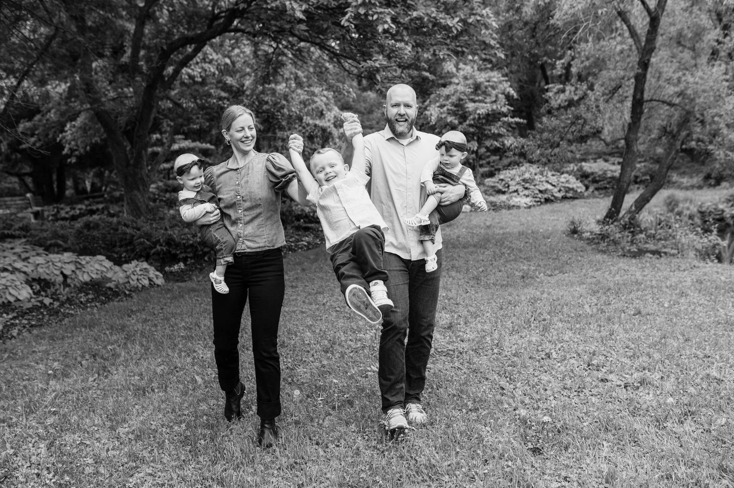 Black and white photo of a happy family of four walking outdoors in a park, with the father holding two young children and the mother holding the hand of another child, all smiling.