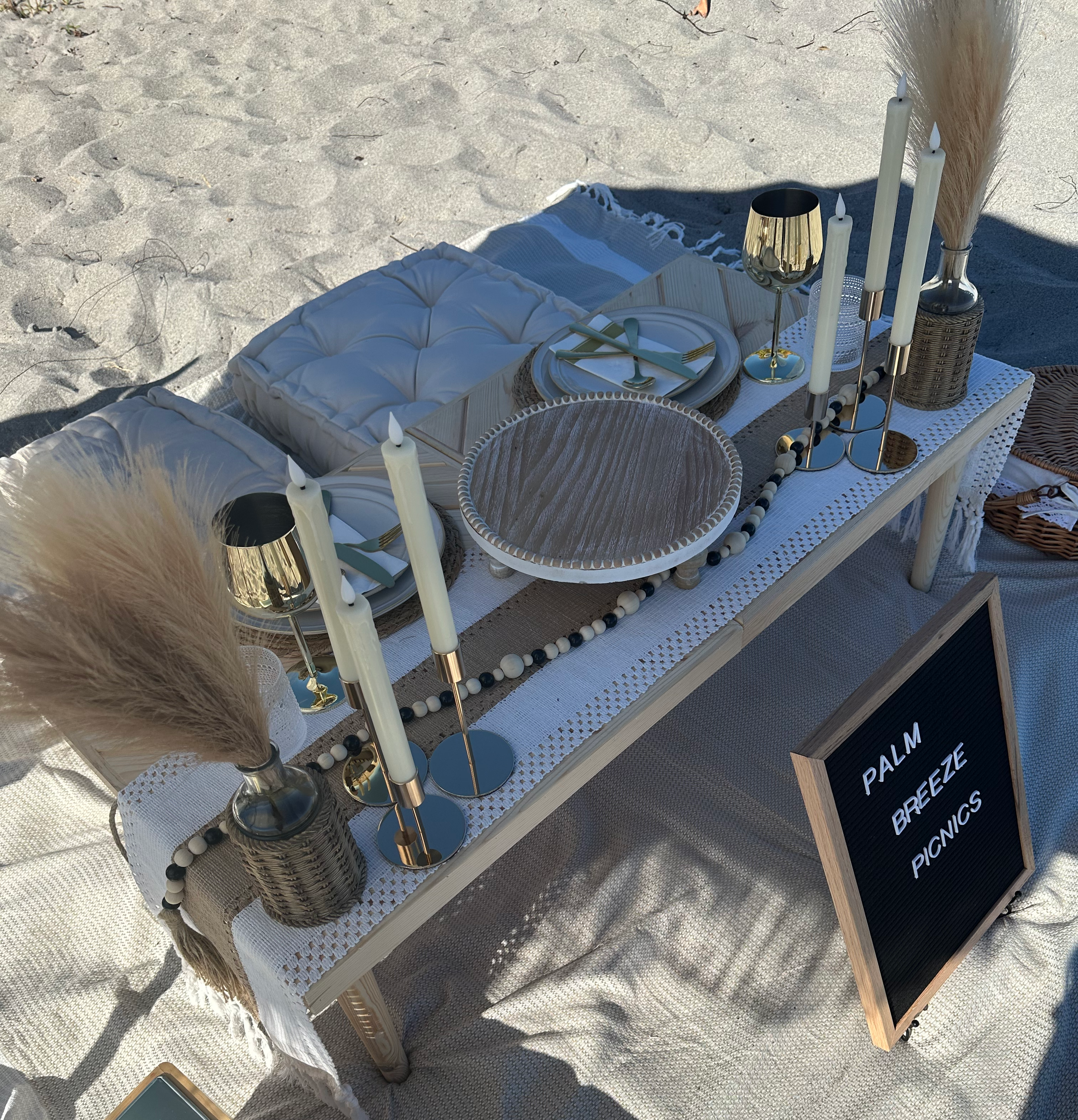 Elegant luxury picnic setup on a Palm Beach County beach with a low wooden table, neutral linens, candles, pampas grass arrangements, gold accents, and floor cushions by Palm Breeze Picnics.