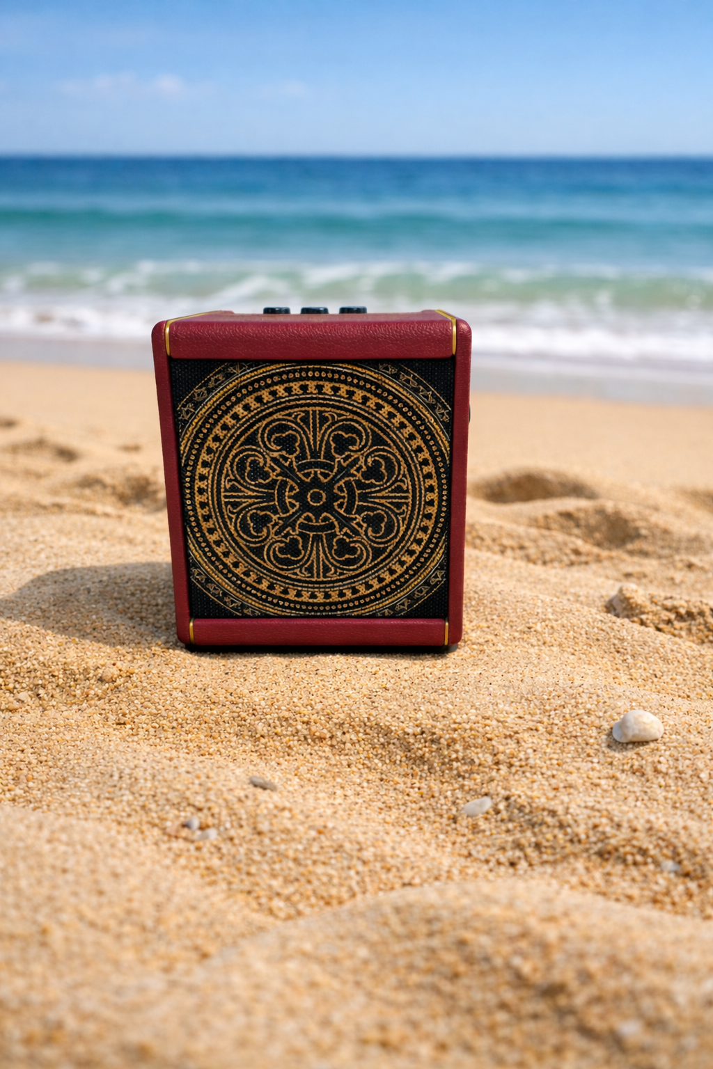 A small amplifier with a decorative black and gold design on the front, placed on sandy beach with ocean waves and blue sky in the background.