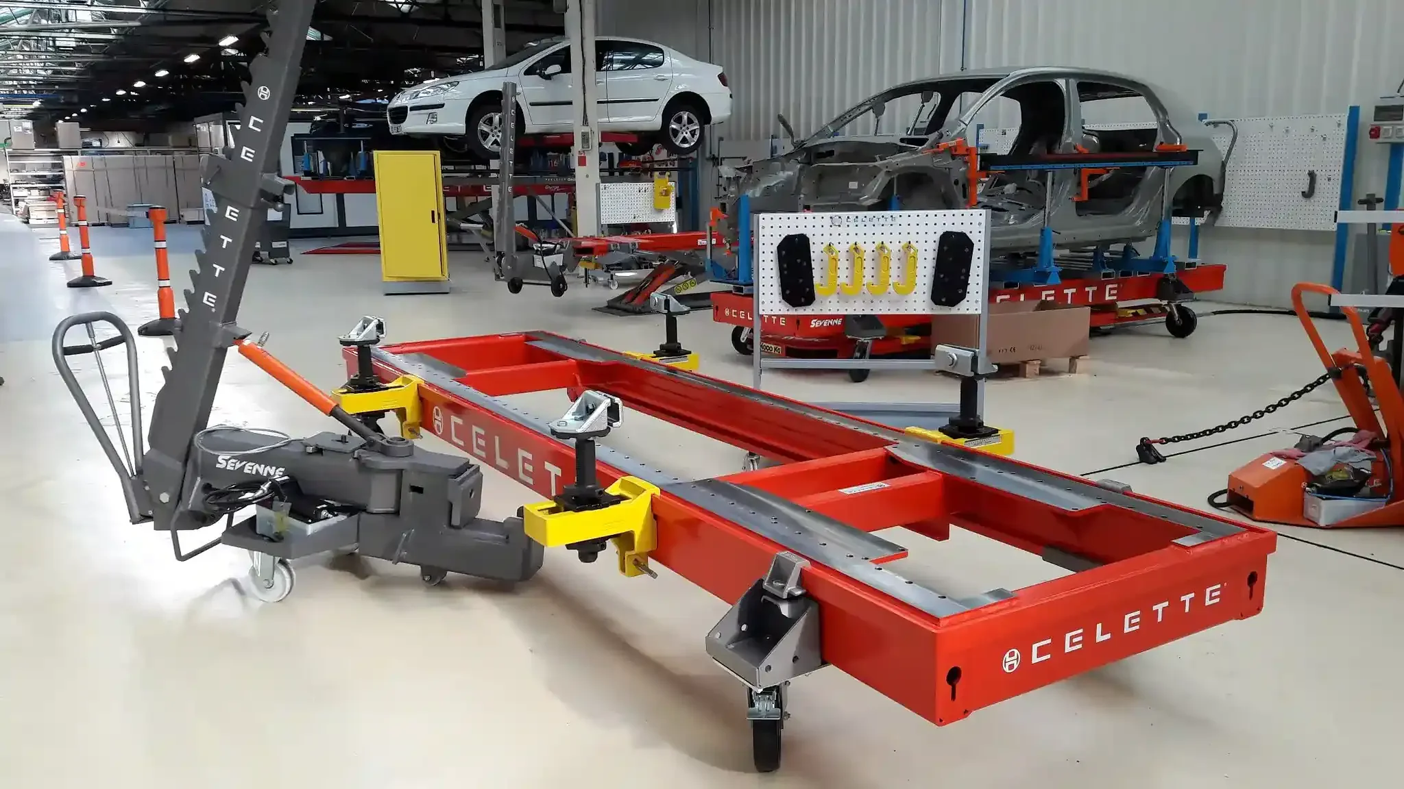 A vehicle assembly and testing facility with various car parts and equipment, including a chassis frame, a partially assembled car body, and car on a lift surrounded by tools and machinery.