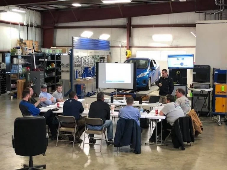A group of people attending a meeting or training session inside an automotive workshop or garage. There's a blue car on a lift in the background, and several screens and equipment around. The attendees are seated around tables, facing the presenter.