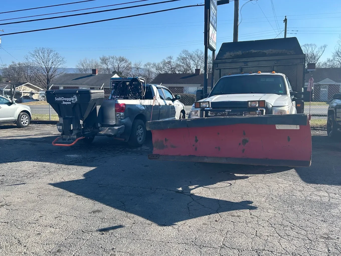 A parking lot with two pickup trucks, one equipped with a snow plow attached to the front, and a third vehicle partially visible on the left. Residential houses and bare trees can be seen in the background.