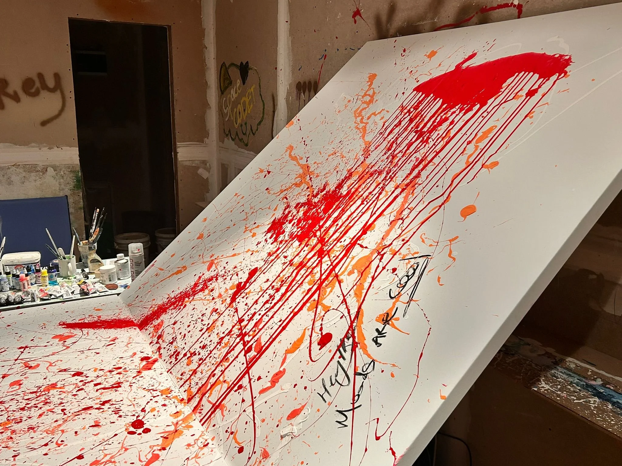 A large white canvas tilted diagonally with orange and red paint splatters and streaks on it. Art supplies like paint bottles and brushes are visible on a table nearby, and the room has unfinished walls with graffiti and artwork.
