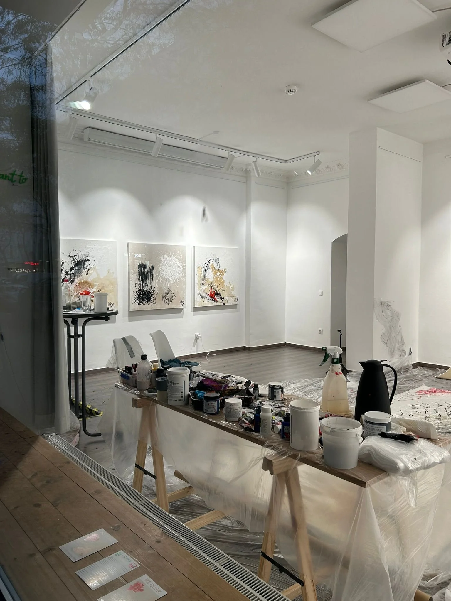 Interior of an art studio or gallery undergoing renovation, with abstract paintings on the wall, art supplies on a table, and a protective plastic cover on the floor.