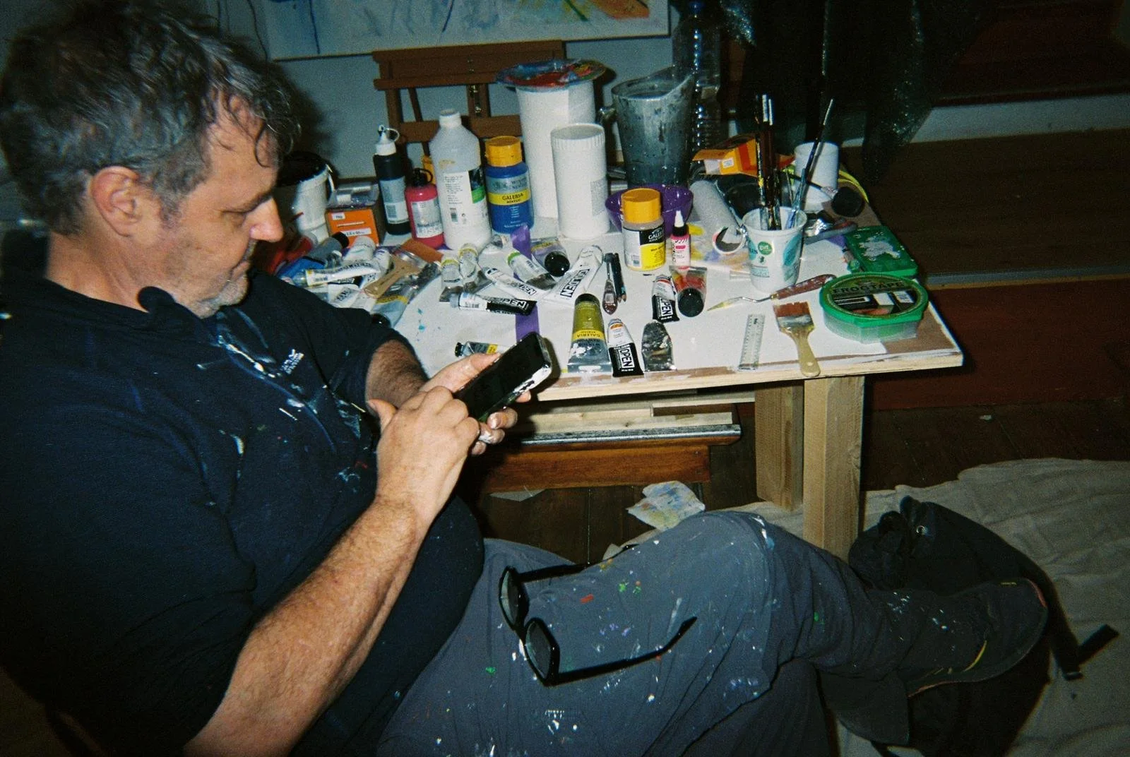 A man sitting at a cluttered table covered with paint supplies such as tubes of paint, brushes, and bottles, while looking at his phone.