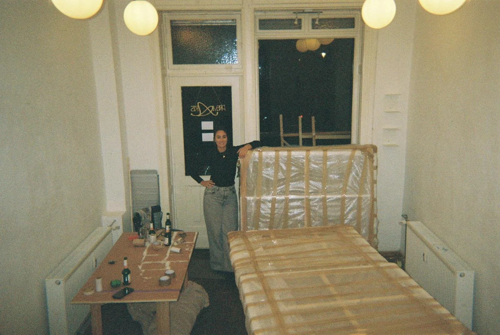 A woman standing in a room with a mattress still in packaging, a small table with bottles and miscellaneous items, and a window behind her. The woman is smiling, wearing a black top and light-colored pants, and has her hand resting on the mattress.