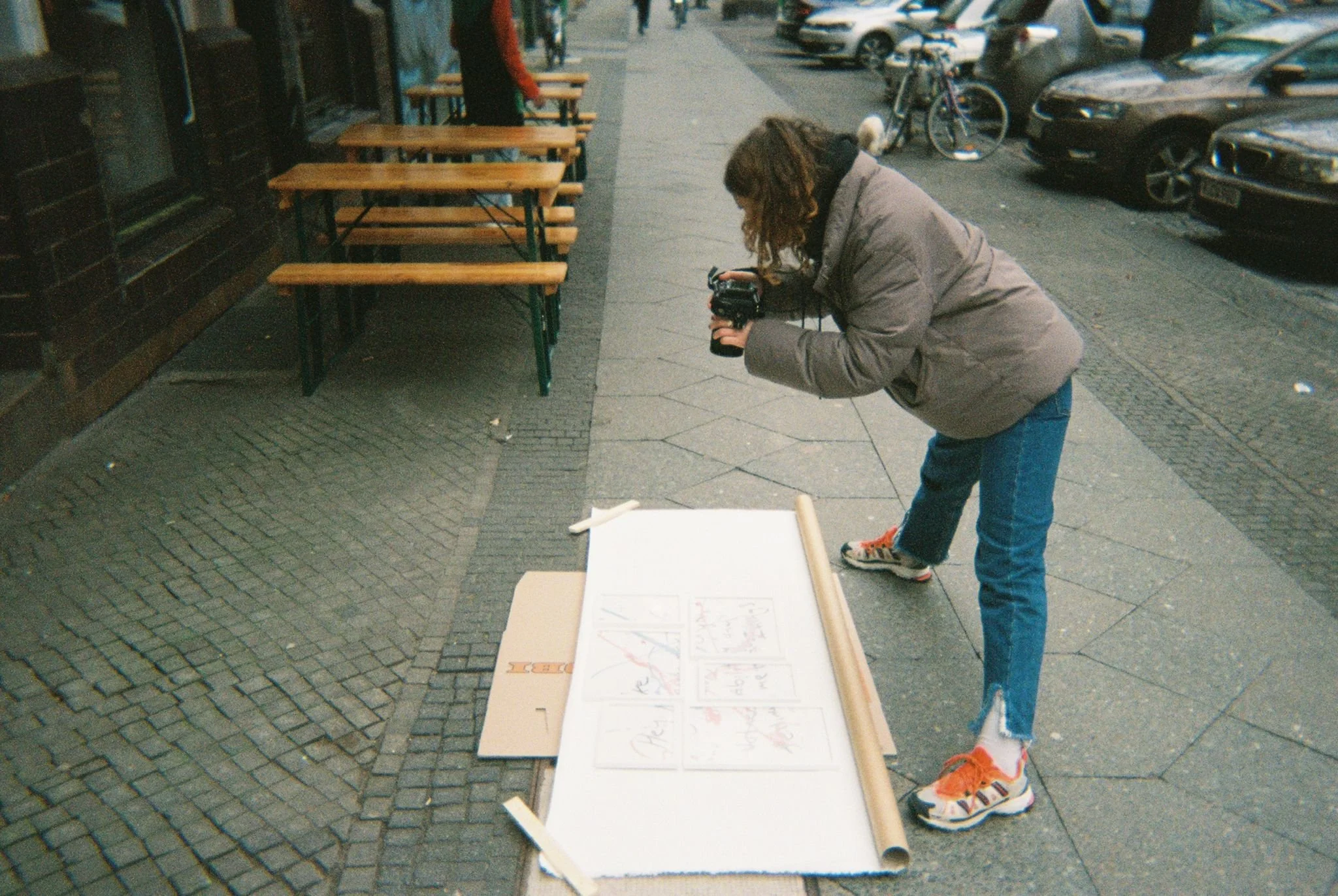 A woman in a gray jacket, jeans, and sneakers is taking a photo of a white poster board with drawings on it on the sidewalk. There are empty outdoor tables and chairs nearby, and parked cars and bicycles along the street.