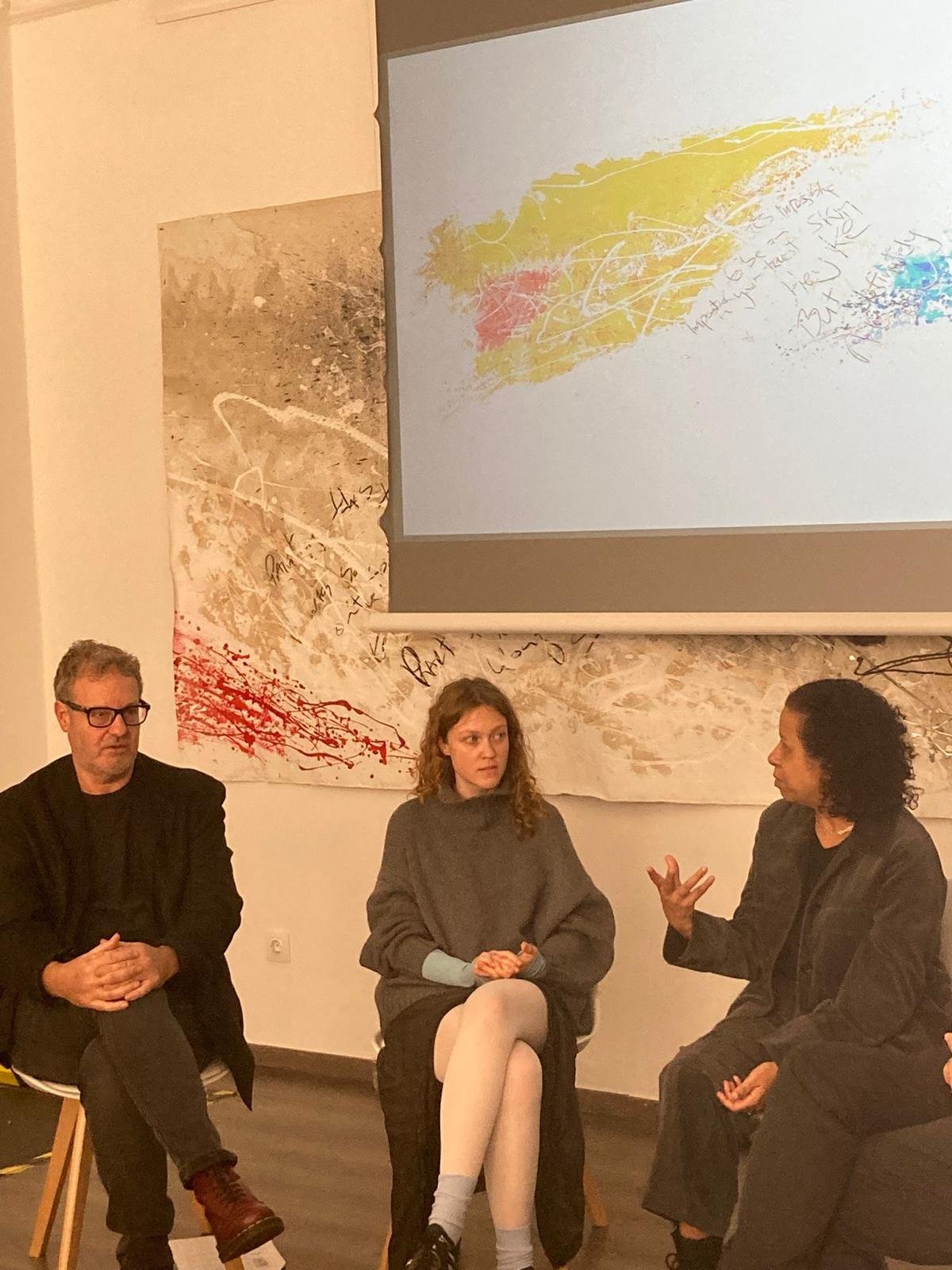 Three people seated in a discussion, two women and one man, in a room with a multicolored, abstract wall decor and a projection screen showing colorful scribbles.