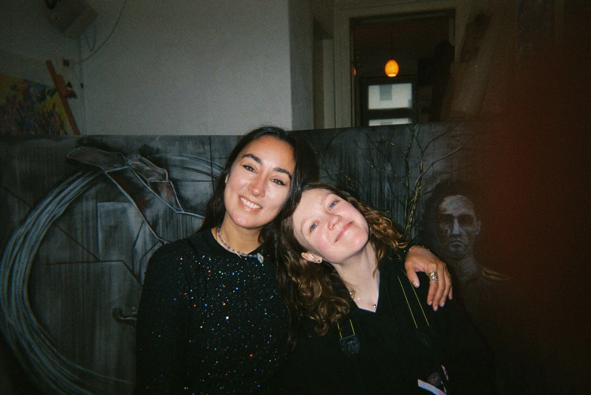 Two women sitting on a couch, smiling, with artwork and a bust sculpture in the background.