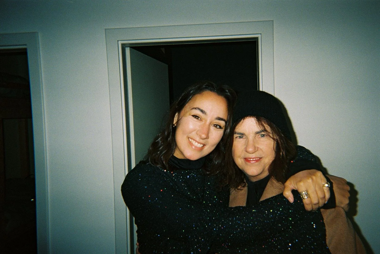 Two women hugging and smiling at the camera, one with dark hair and the other with brown hair and wearing a black beanie, indoors with white walls.