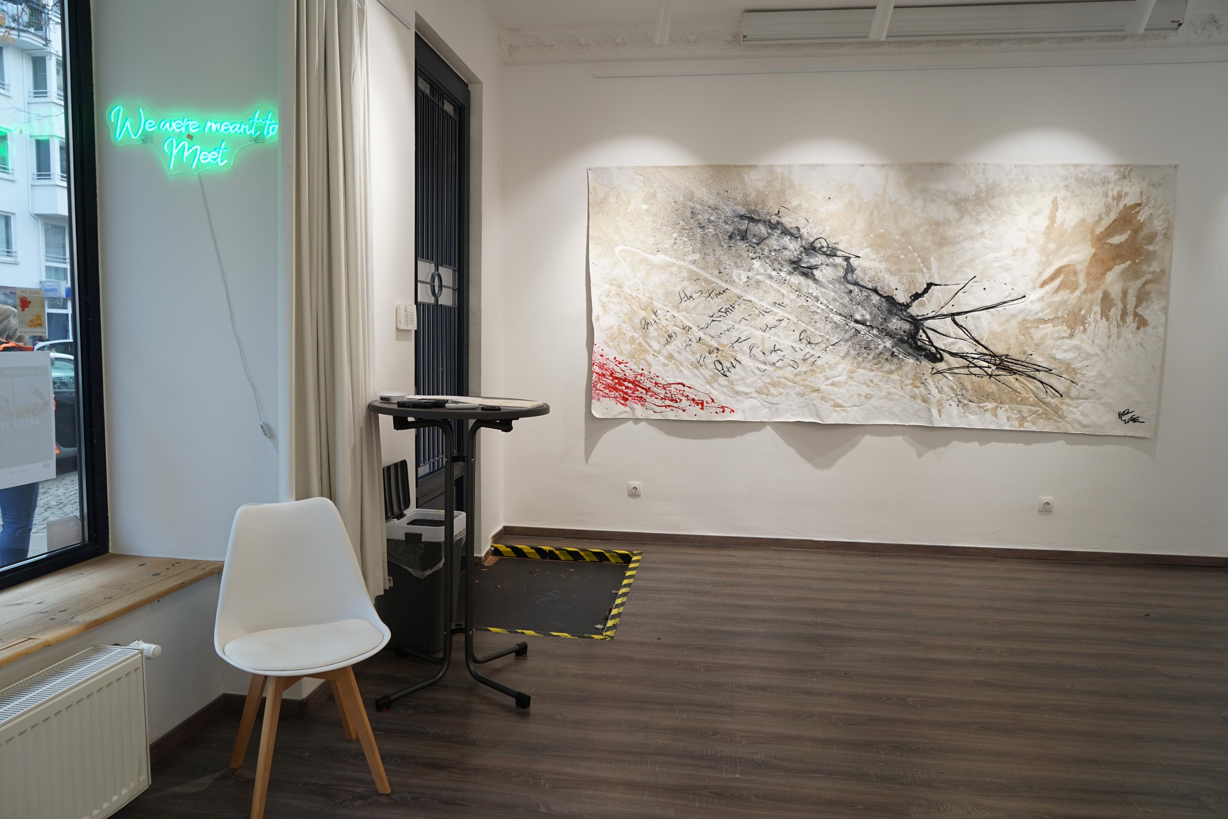Interior of an art gallery with a large abstract painting on the wall, a white chair near the window, and a neon sign that reads 'We were meant to meet' on the wall.