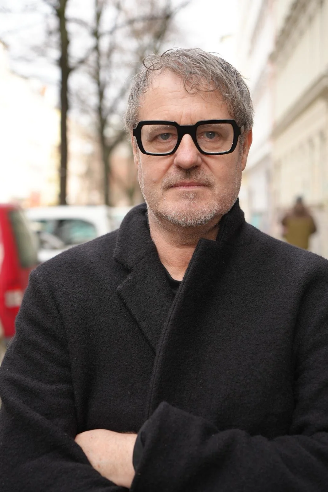 A middle-aged man with gray hair and beard wearing large black glasses and a black coat, standing outdoors on a street with cars and trees in the background.