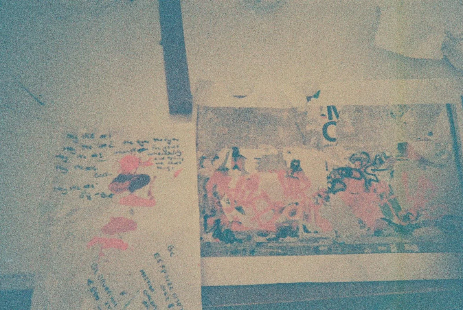 Photograph of two handwritten notes and a colorful artwork on a wall. One note has text and a cartoon image of a person with hair, the other note and artwork are partially visible.