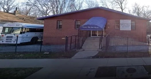 A brick building with blue awning and steps leading to the entrance, fenced yard in front, and a school bus parked nearby.