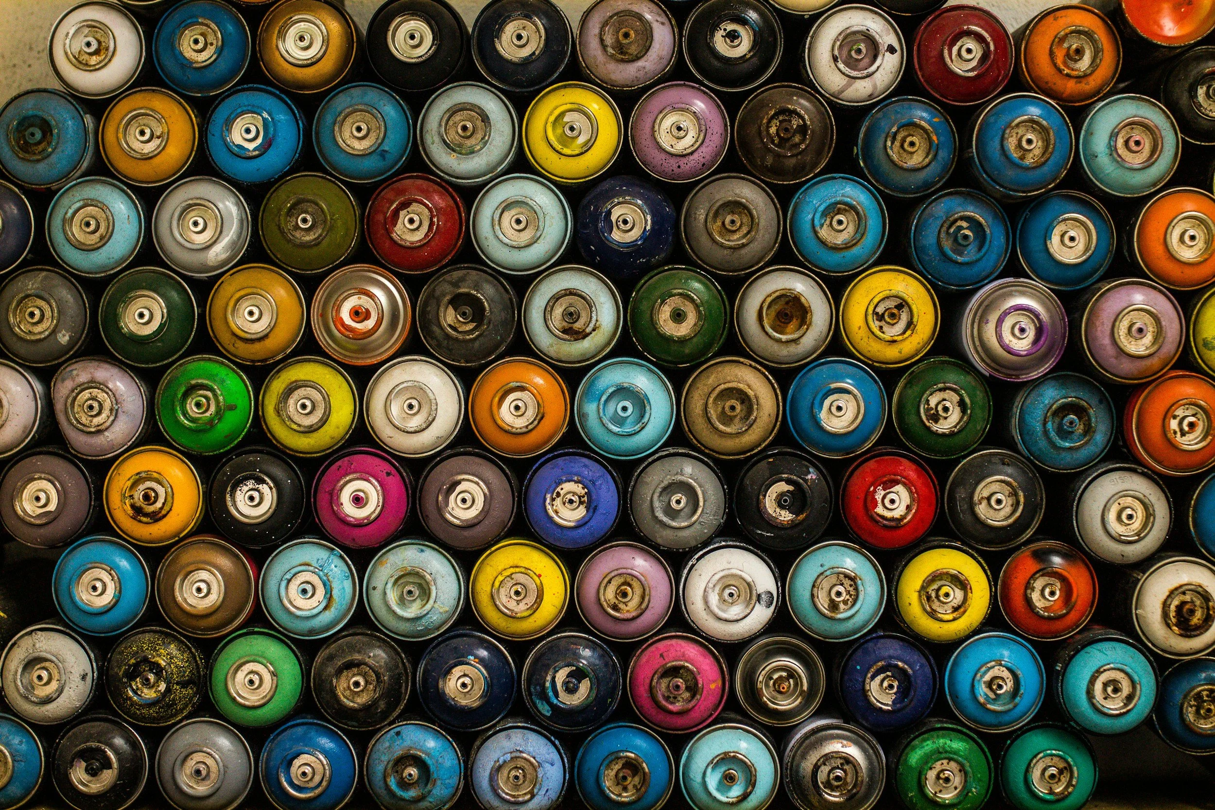 Top-down view of spray paint cans arranged in rows, featuring various colors including blue, yellow, green, red, purple, black, white, and orange, with visible nozzles and caps.