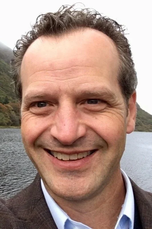 Close-up of a smiling man with curly hair in an outdoor setting near water and trees in the background.