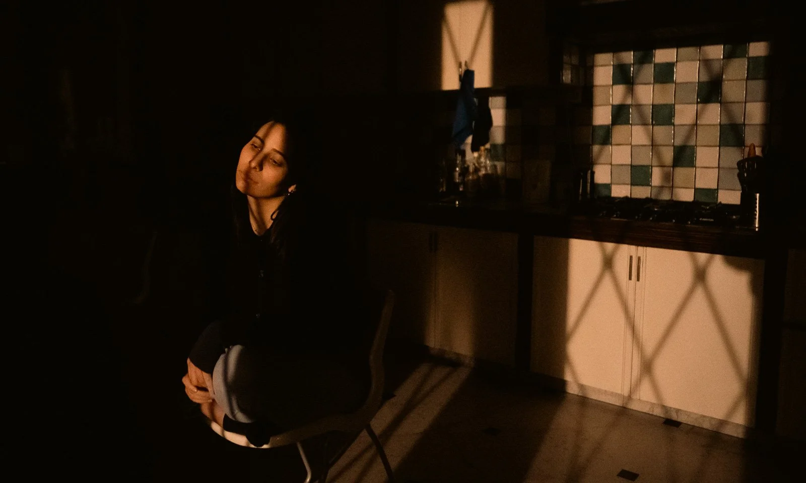 A girl sits in her kitchen at night, appearing sad as the light filters through the windows