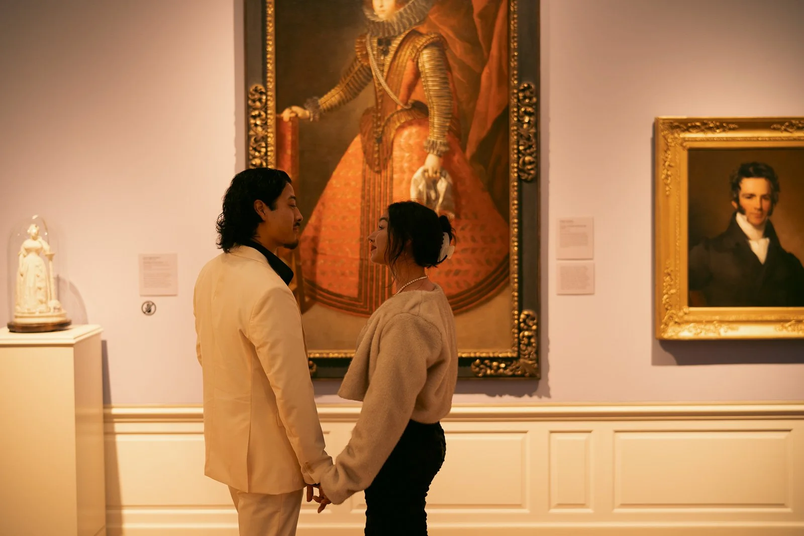  A couple in matching white outfits stand in front of large paintings smiling at each other 