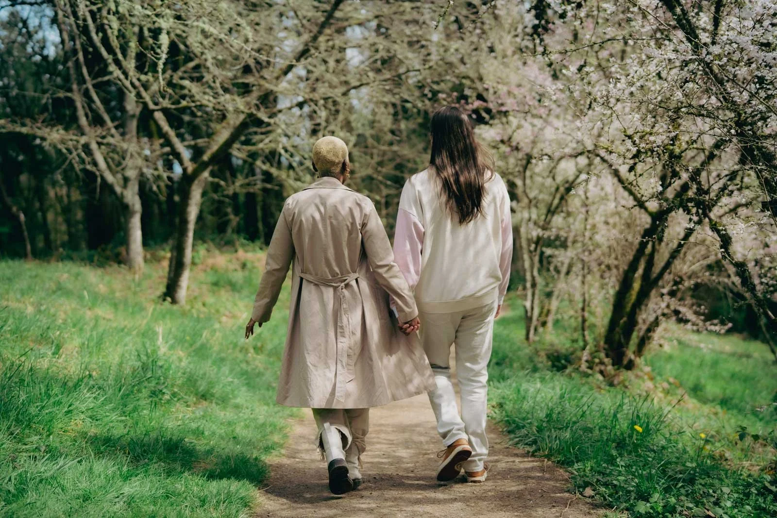  A couple walks down a forest trail holding hands with flowering cherry blossom trees on either side 
