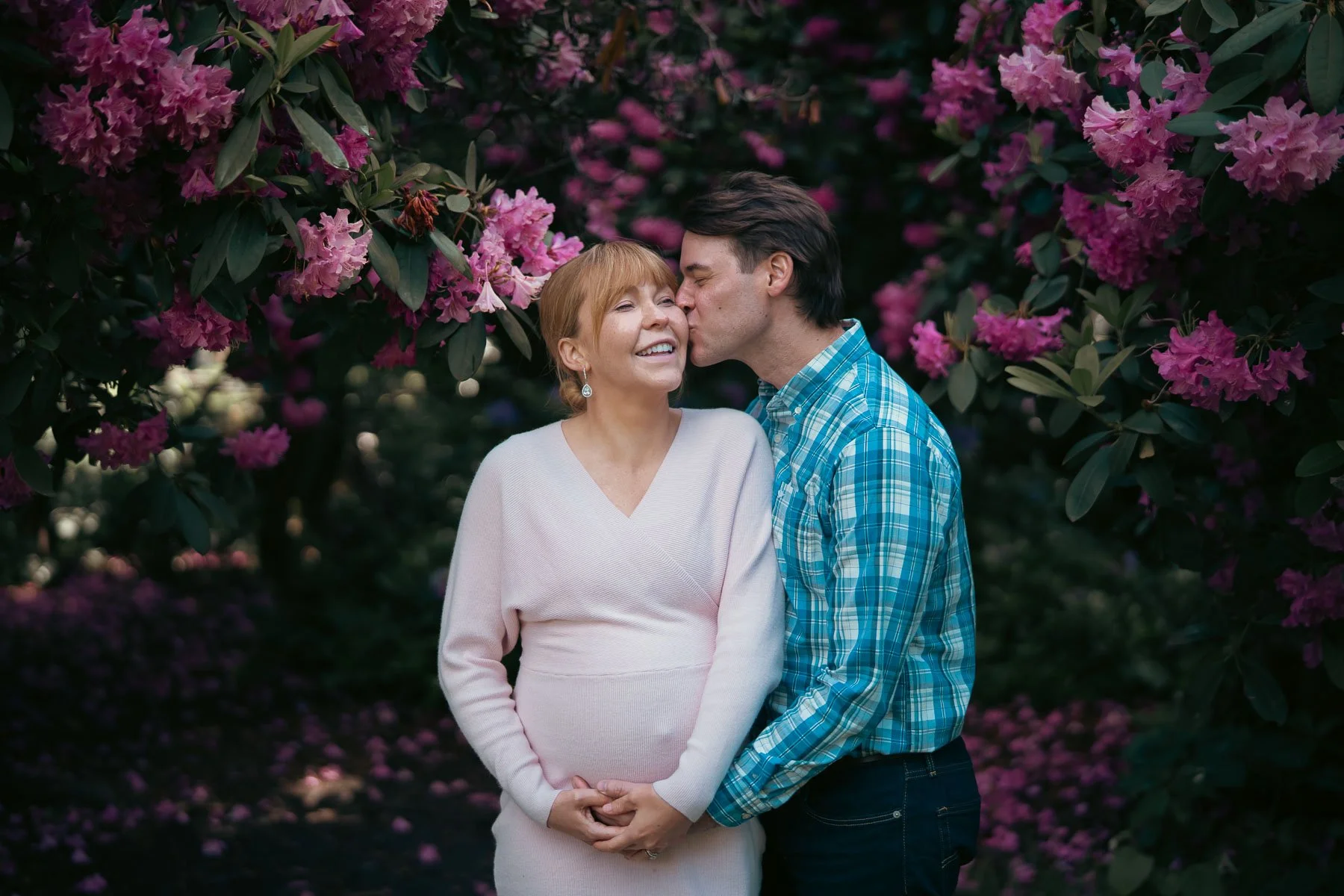 A smiling pregnant woman with her husband holding her hand and kissing her check in front of a flowering tree