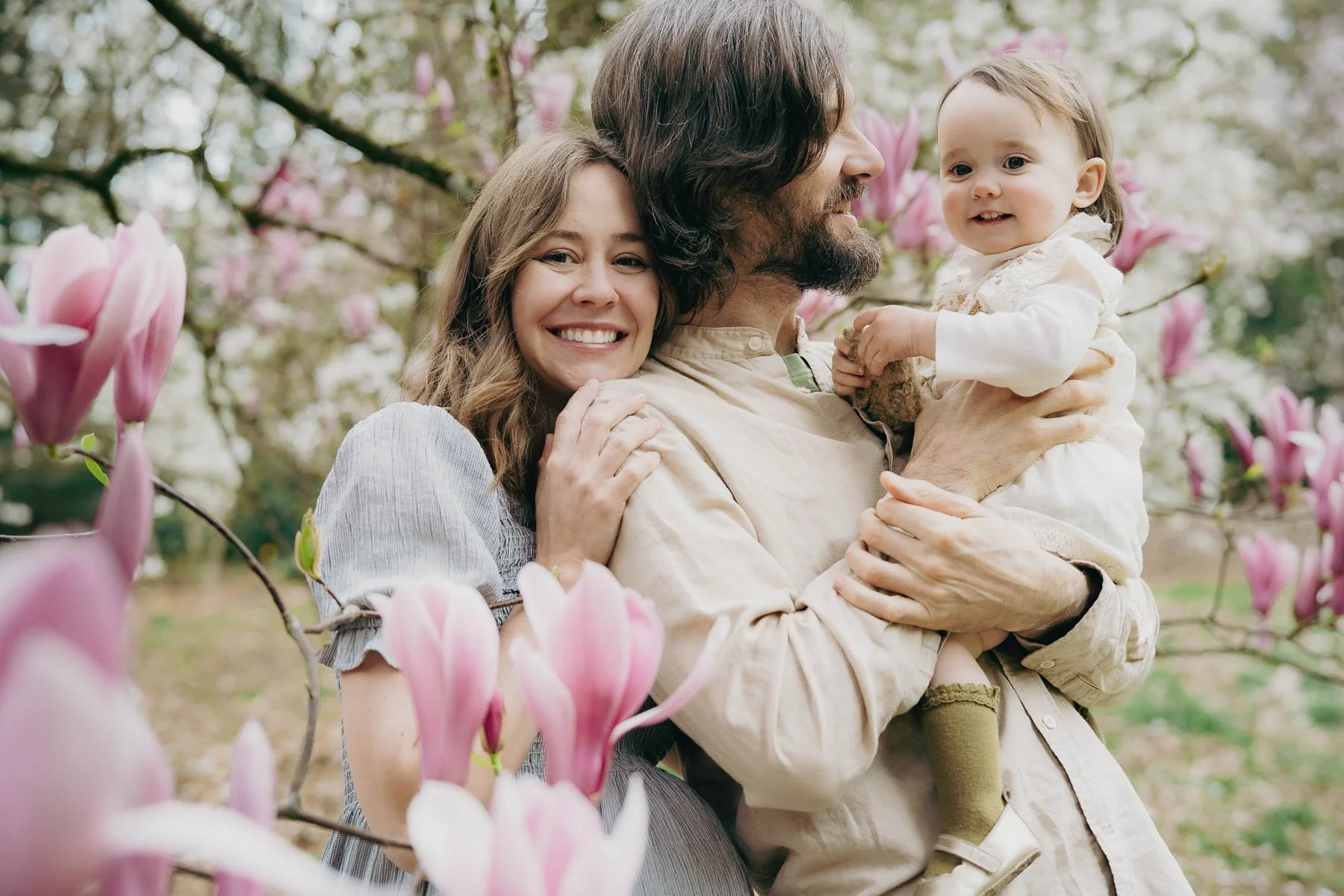 A pregnant mother, her husband and child all smiling and hugging each other in a field of flowering Magnolia trees