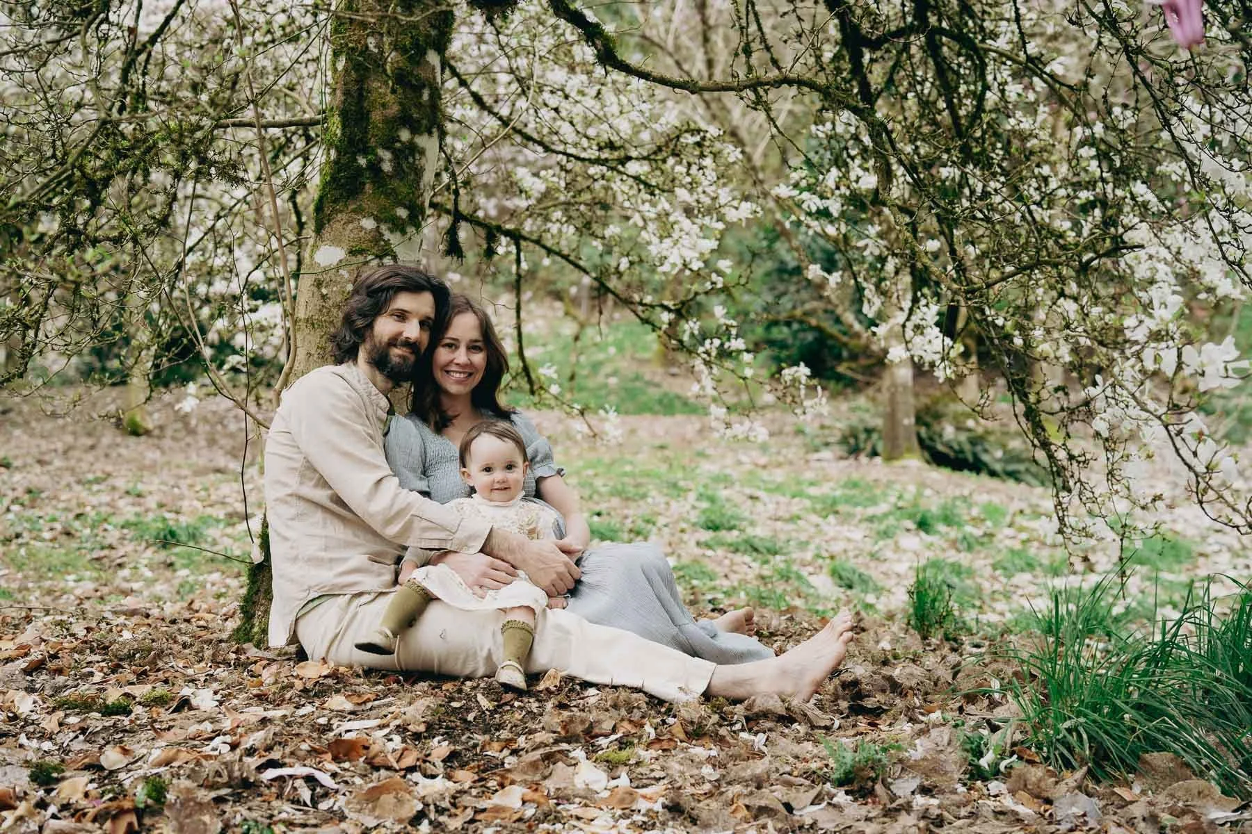 A smiling family dressed in light earth tones sit under and blossoming tree in Hoyt Arboretum