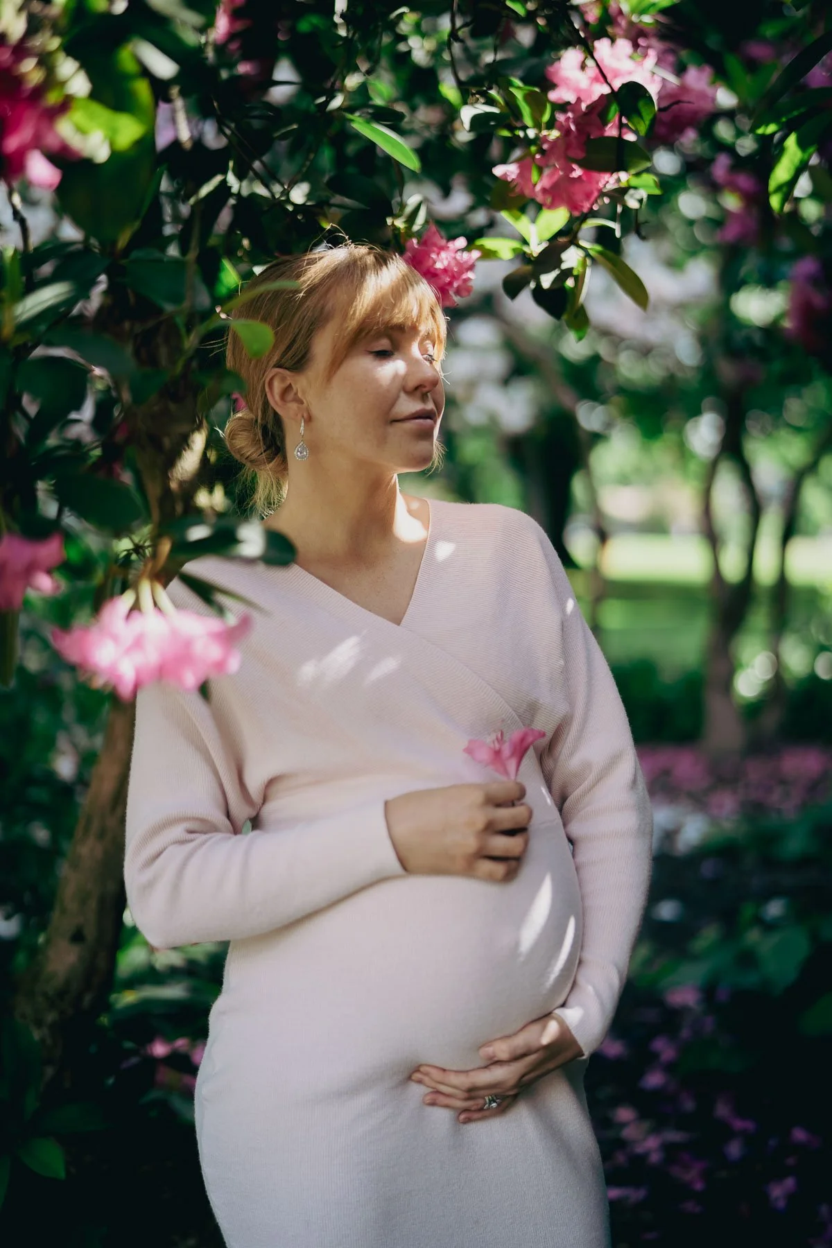 A pregnant woman with her eyes closed holds her stomach while rays of light fall on her face through the flowered branches around her