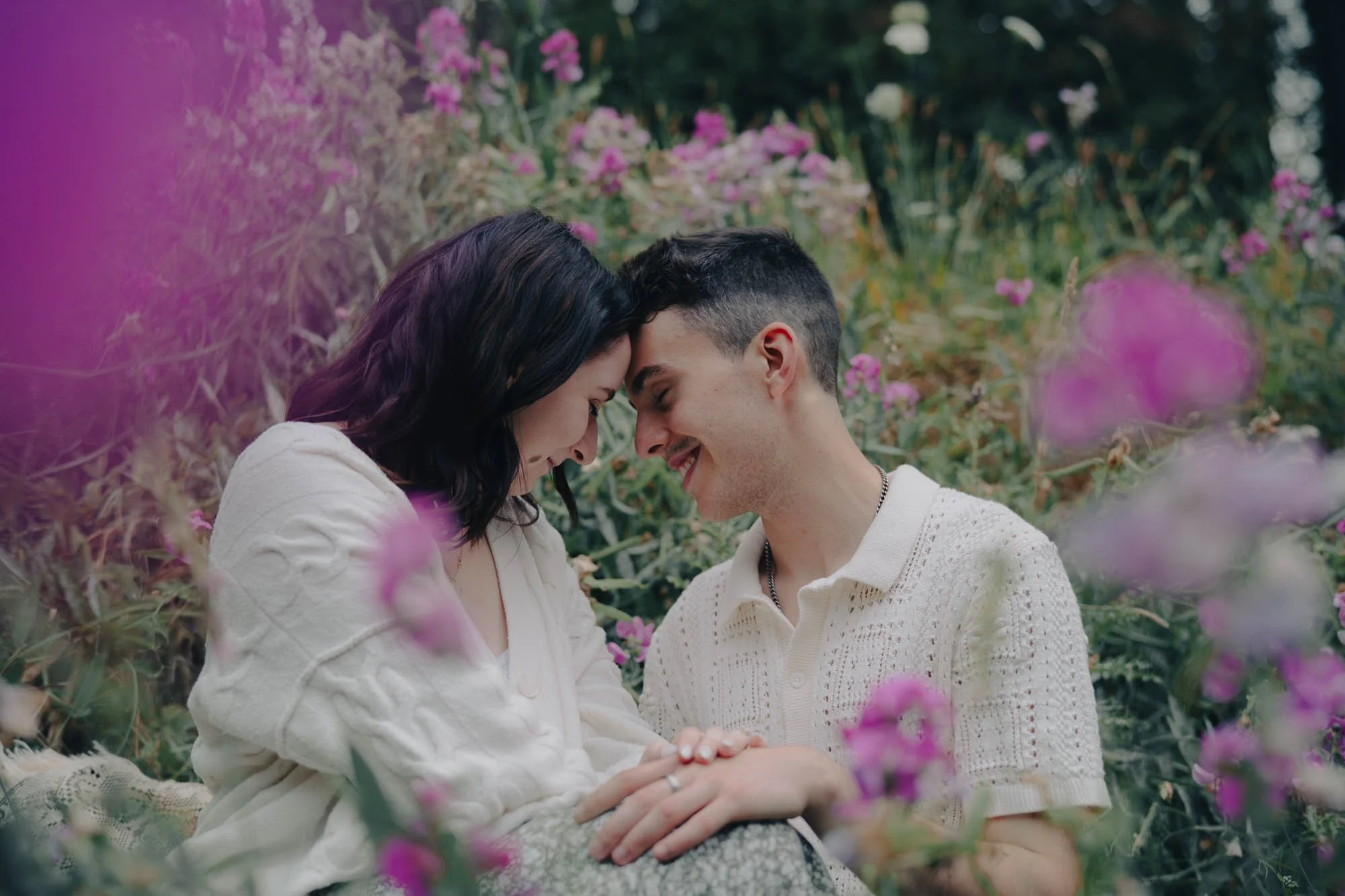 A couple in matching white outfits lean their foreheads together framed by a field of purple flowers