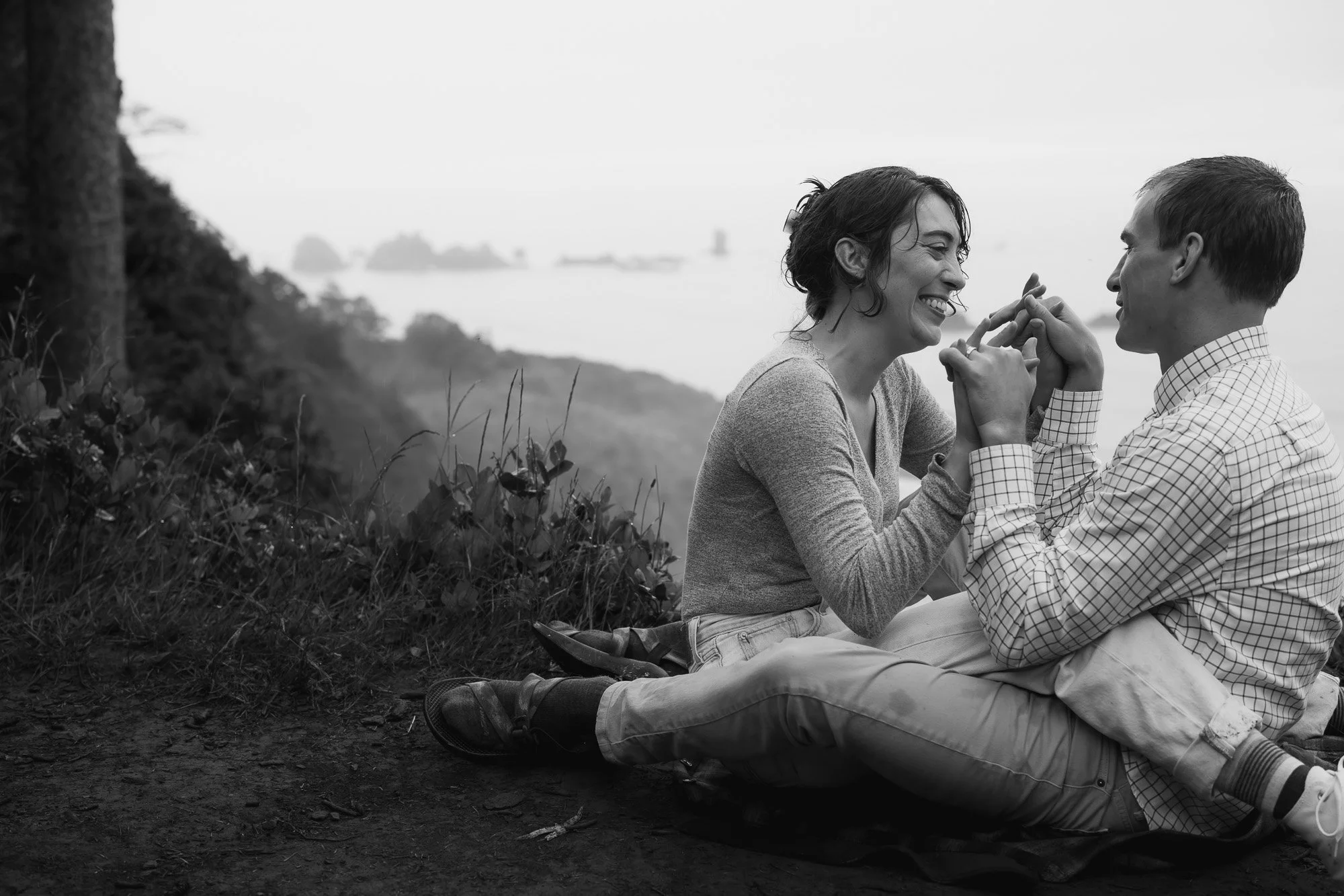 A smiling couple sits entangled at the edge of a cliff looking over Indian Beach
