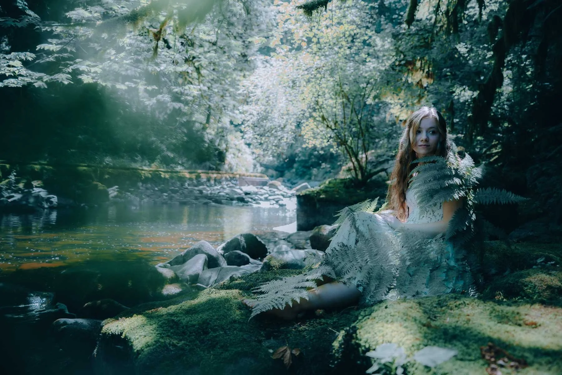 Girl sitting for a senior portrait at the edge of a river with ferns covering her