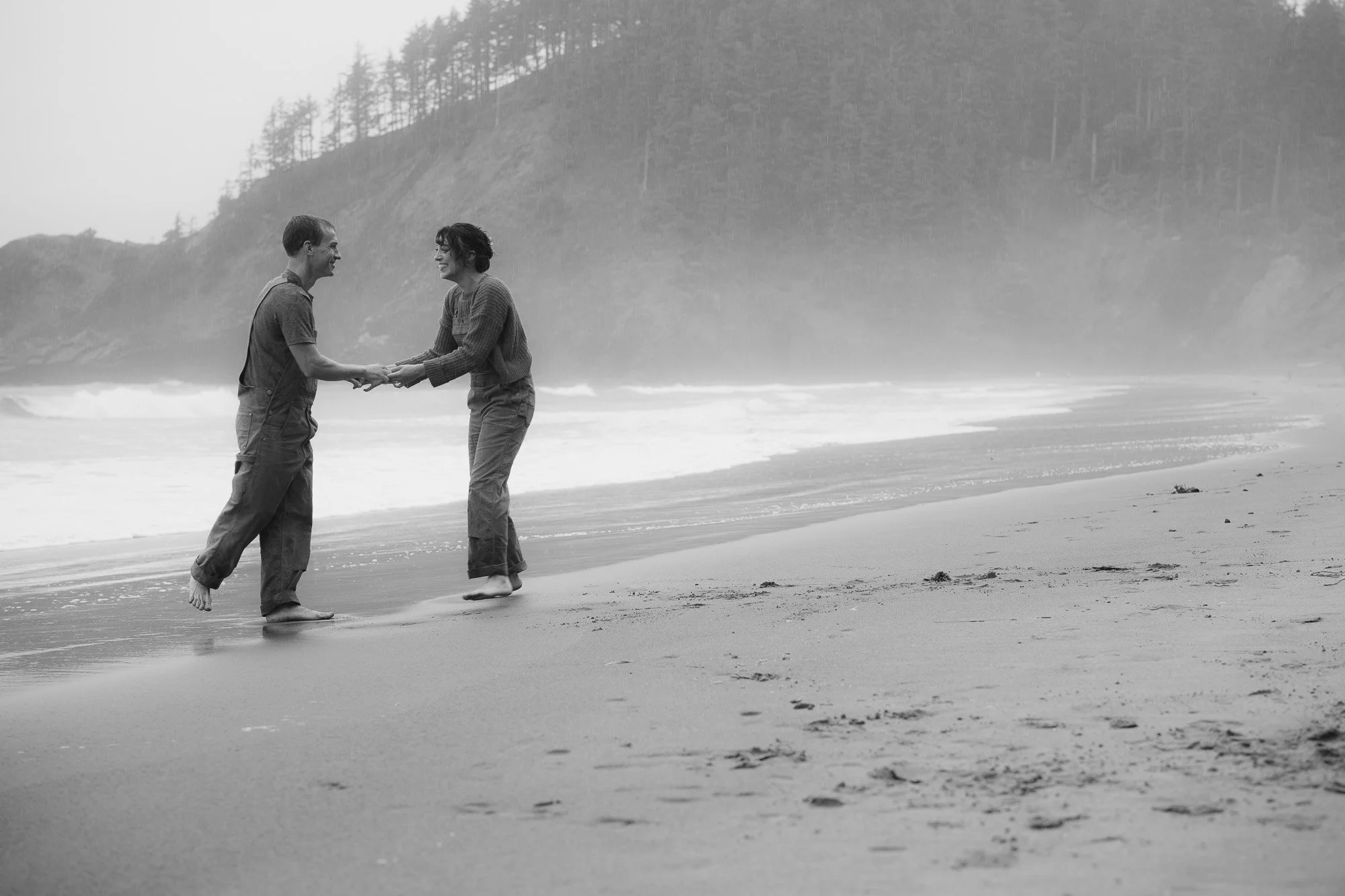 A couple playfully dancing on the beach with misty cliffs behind them