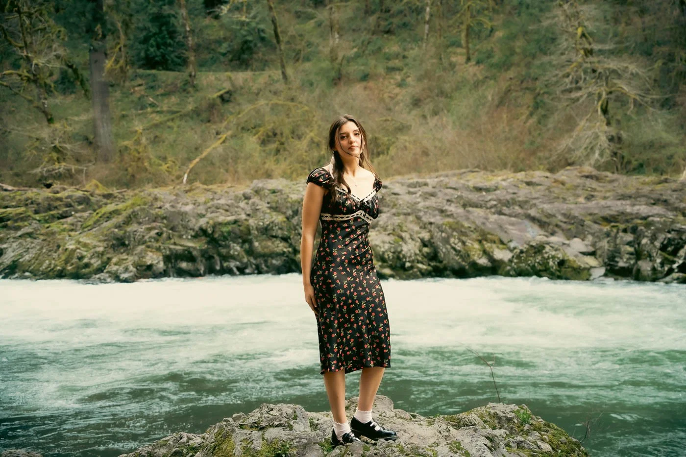 A girl in a black dress standing at the edge of a rapid river