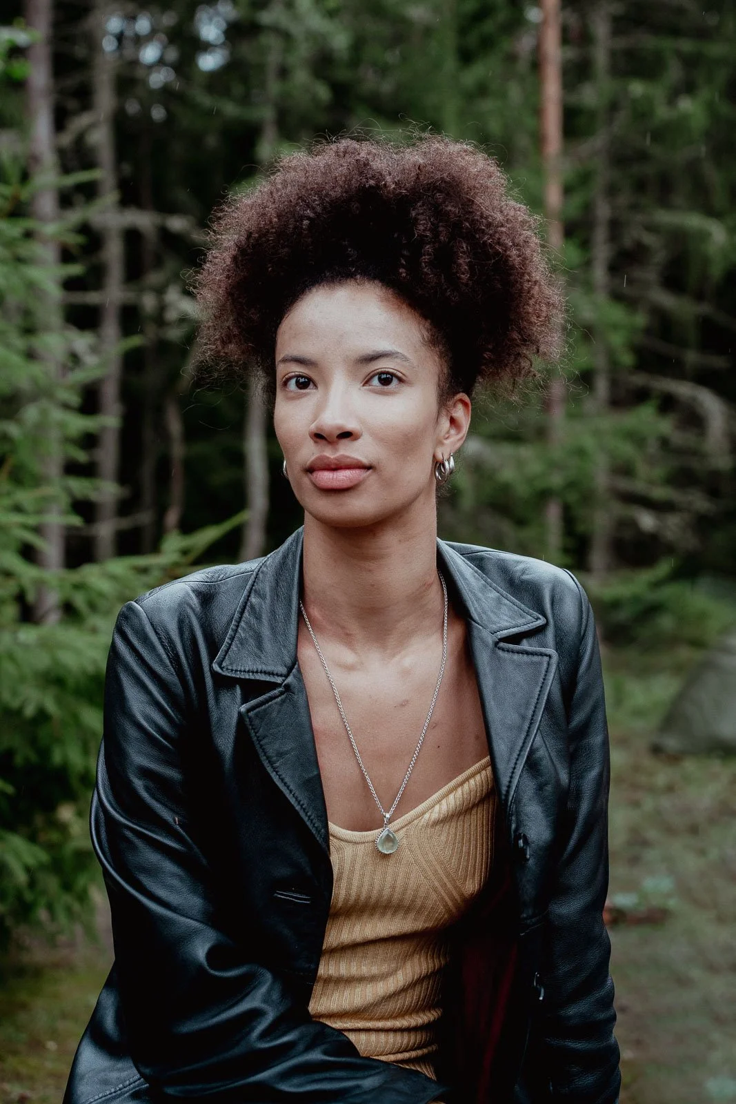 An attractive black woman in a leather jacket with the forest behind her