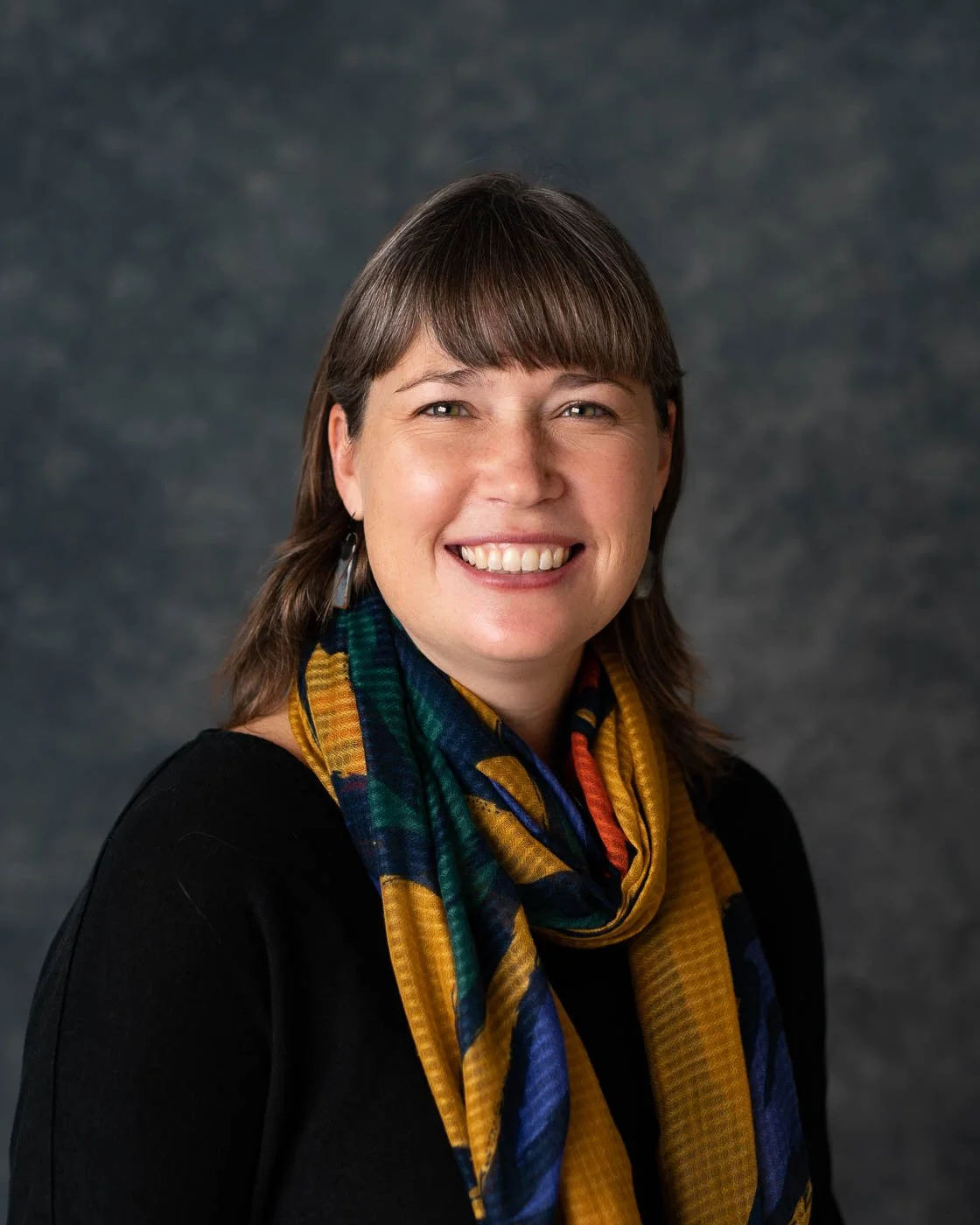 Headshot of a smiling woman wearing a multi-colored scarf against a mottled grey background