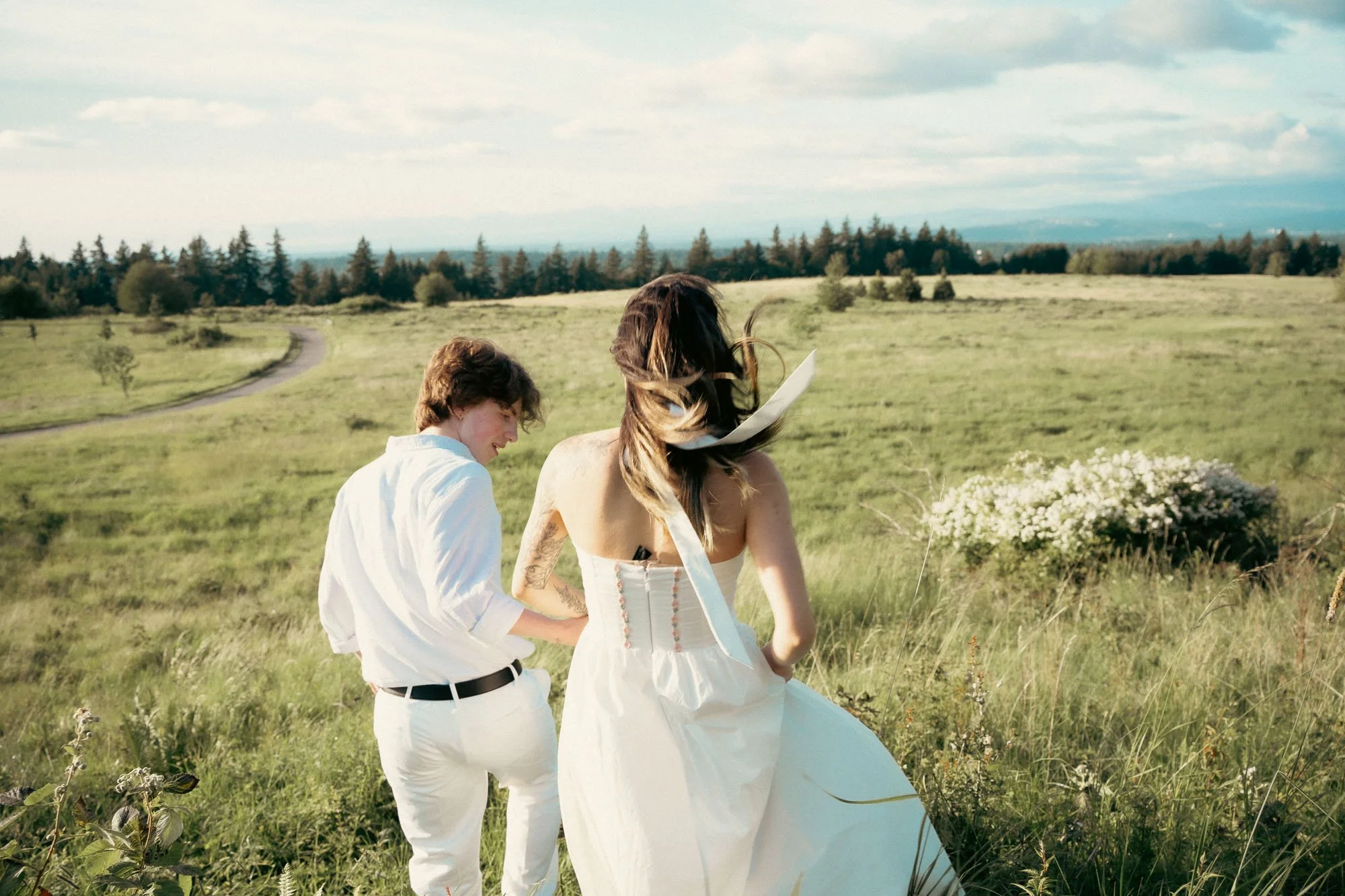 A couple dressed in white walks down a grassy hill into a meadow