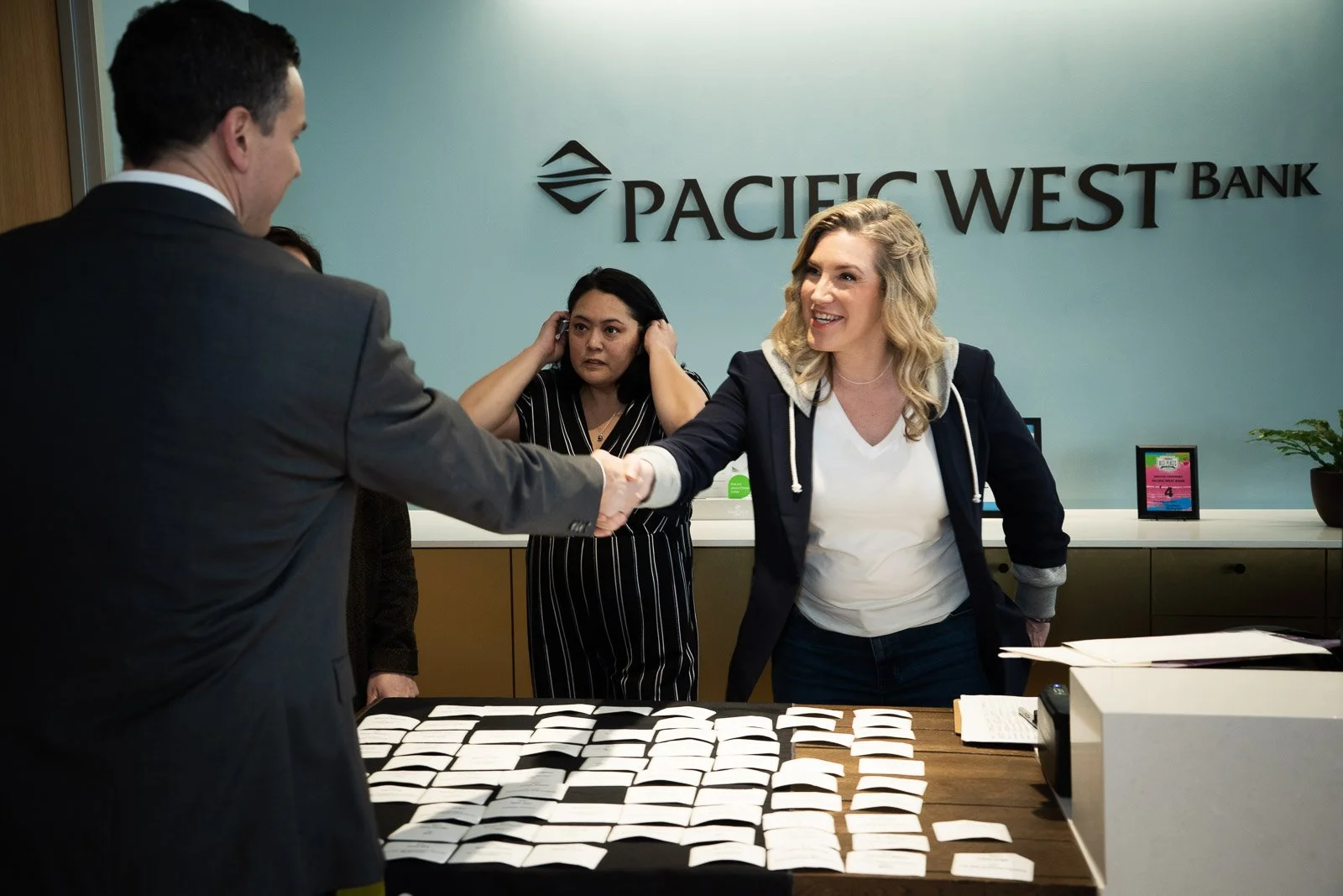A woman shaking hands and smiling in front of a Pacific West Bank sign
