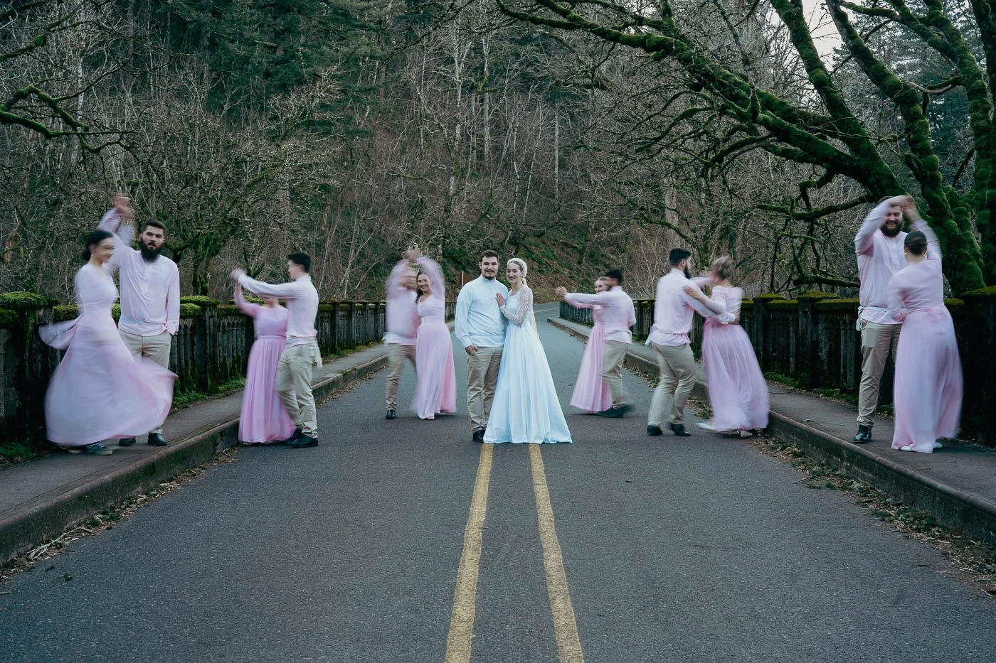 Wedding couple posed on a quiet, tree-lined street with friends dancing and celebrating around them.