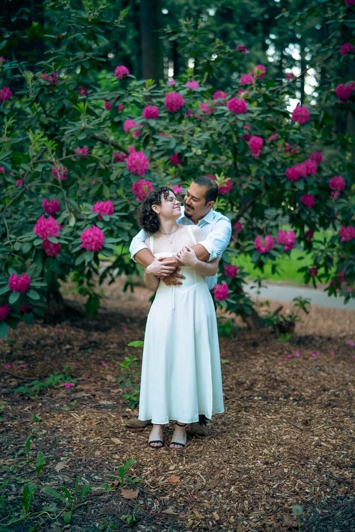 A couple dressed in white standing in front of a flowering Rhododendron tree