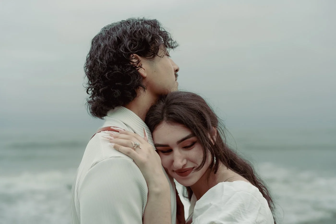 A smiling woman leans into a mans chest with the ocean behind them