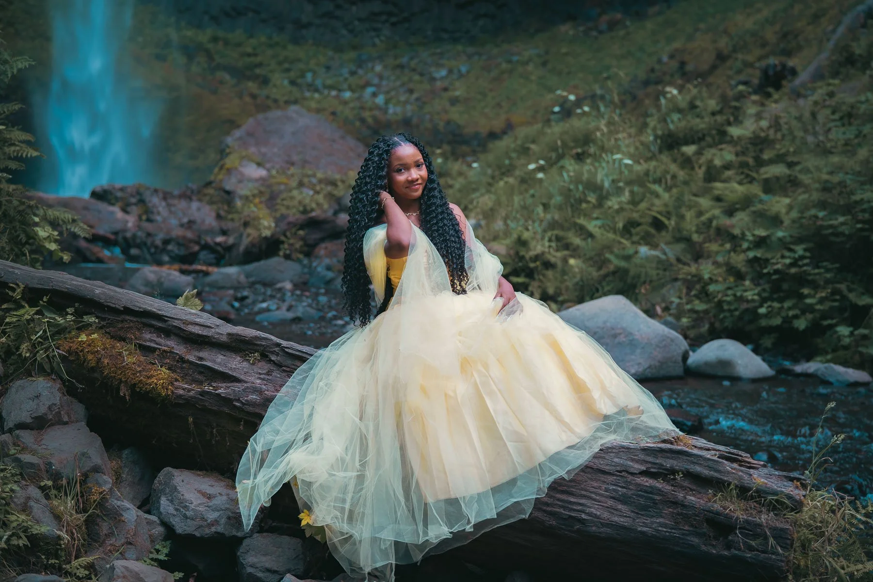 A high school senior in a yellow dress sitting on a log in the river with a waterfall