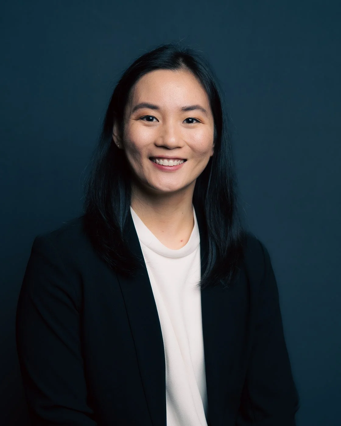 Studio headshot of a young woman in a black blazer and white shirt set against a dark blue background