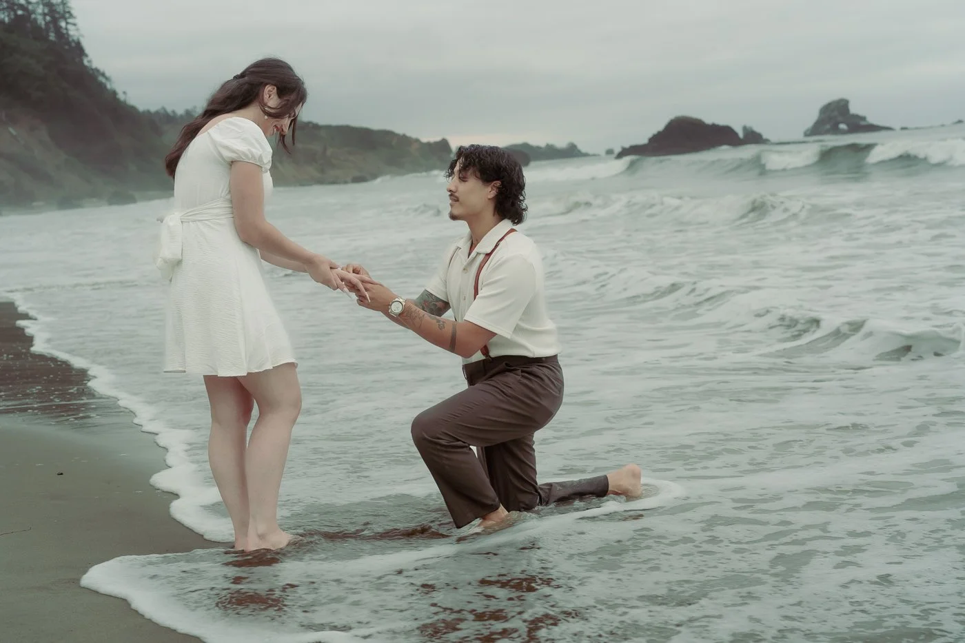 A man knees in the ocean proposing to a woman dressed in a white skirt