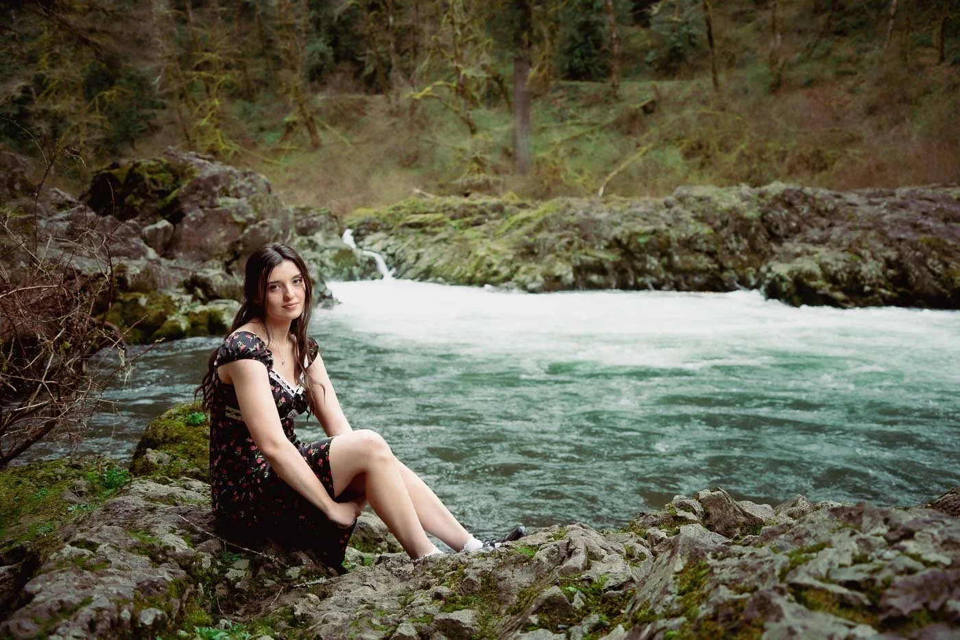 Senior portrait of a girl in a black dress sitting at the rivers edge