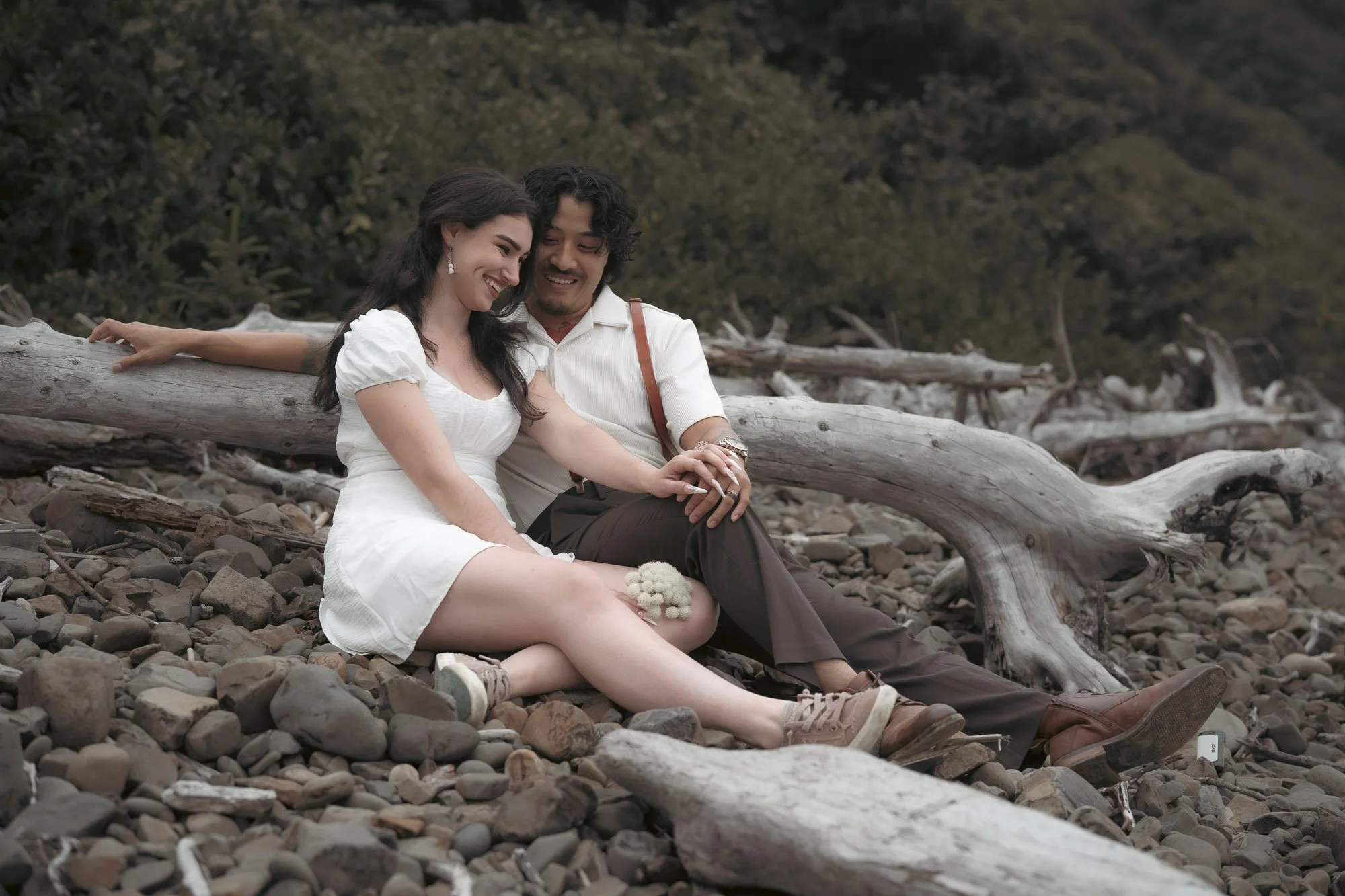 An engaged couple sits on a rocky beach, holding a bouquet of wild flowers and looking at the engagement ring
