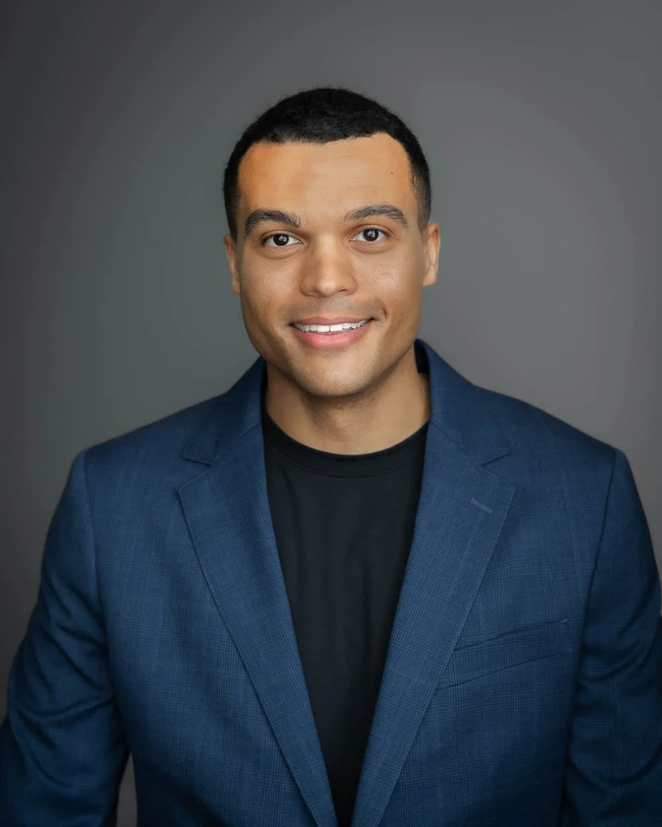 Headshot of a young black man wearing a black shirt and blue blazer.