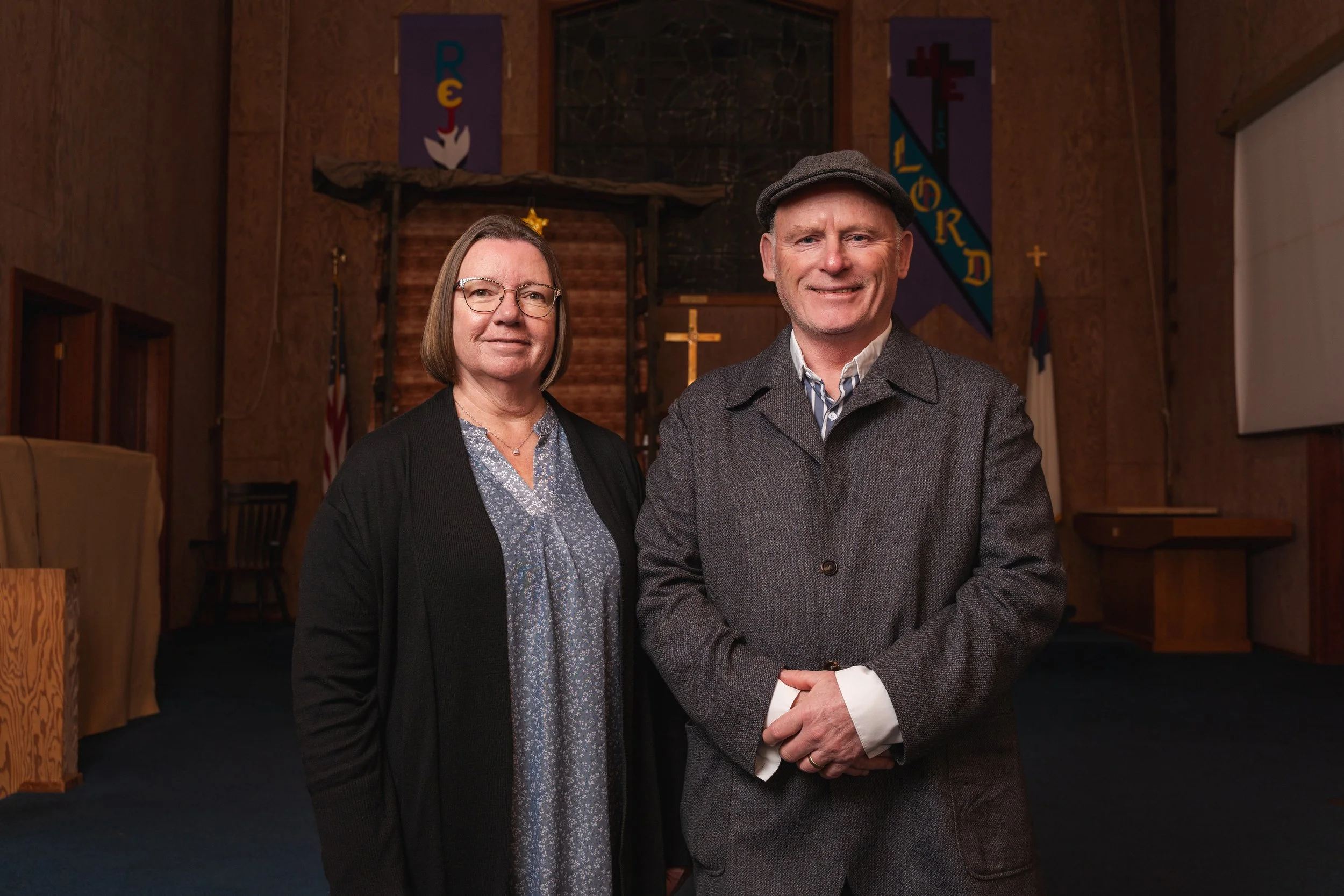 A middle-aged woman and man standing inside a church with religious banners and a cross behind them. The woman has shoulder-length hair, glasses, and is wearing a patterned blouse with a dark cardigan. The man is wearing a gray coat, striped shirt, and a cap, smiling at the camera.