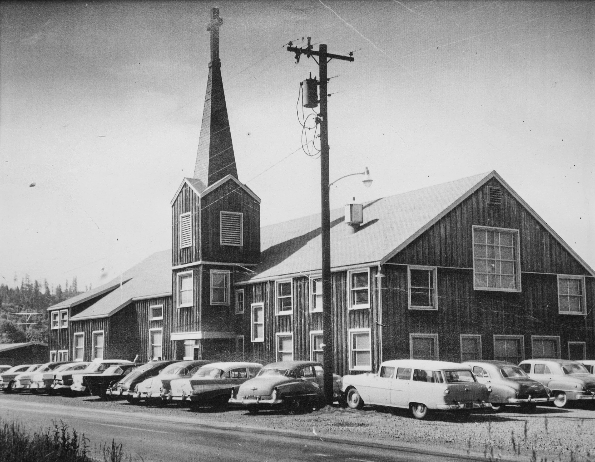 Black and white photo of a vintage wooden church with a tall steeple and large front window, situated next to a parking lot filled with classic cars.