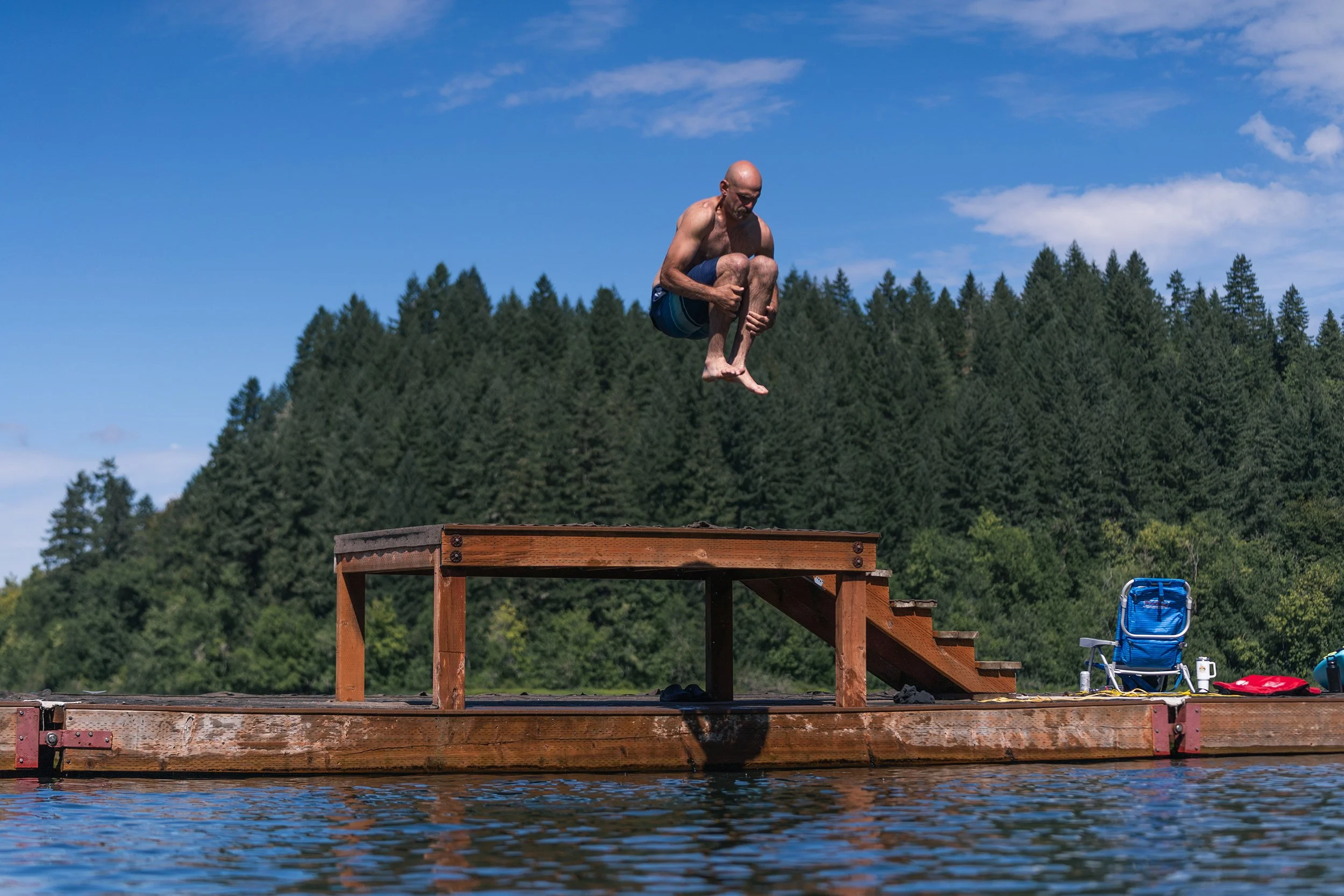 A man jumping off a wooden dock into a lake on a clear day with a forest in the background.